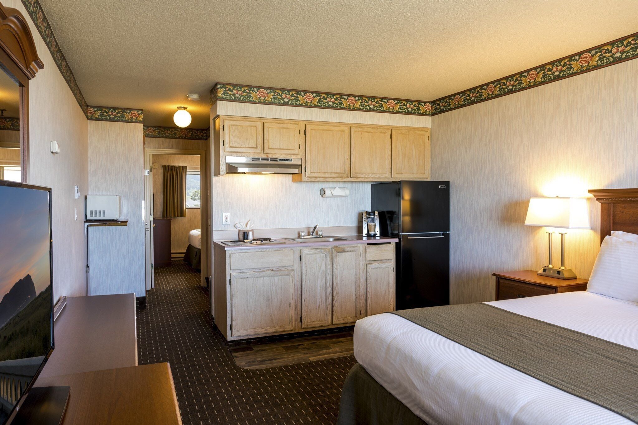 A compact hotel room with a kitchenette, a bed, a desk, and a television.
