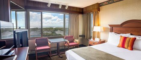 Ein Hotelzimmer mit einem großen Bett, einem Schreibtisch, einem Sessel, einem Fernseher und Blick auf den Strand.