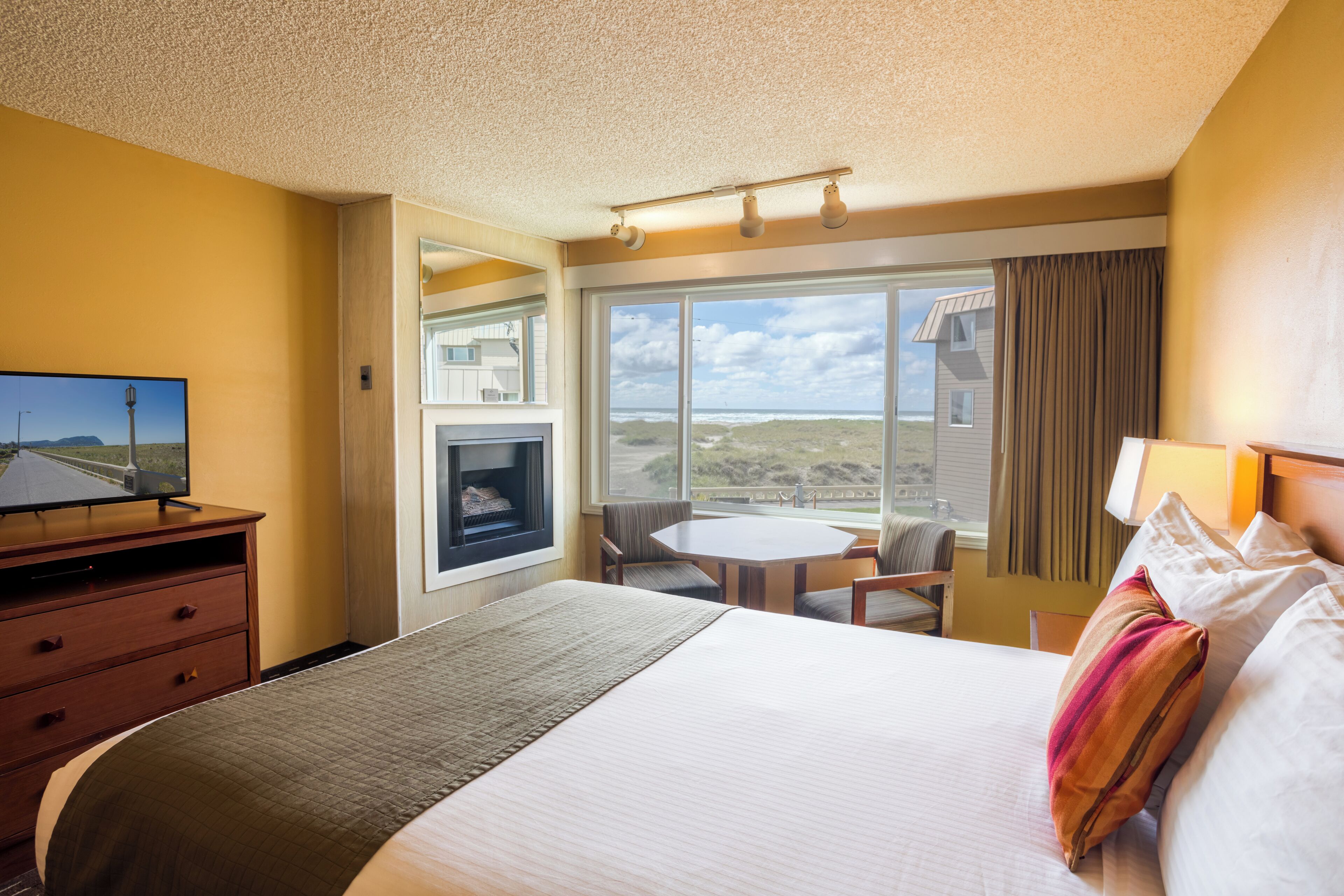 Quarto de hotel com uma cama grande, televisão, mesa de jantar e vista para a praia.