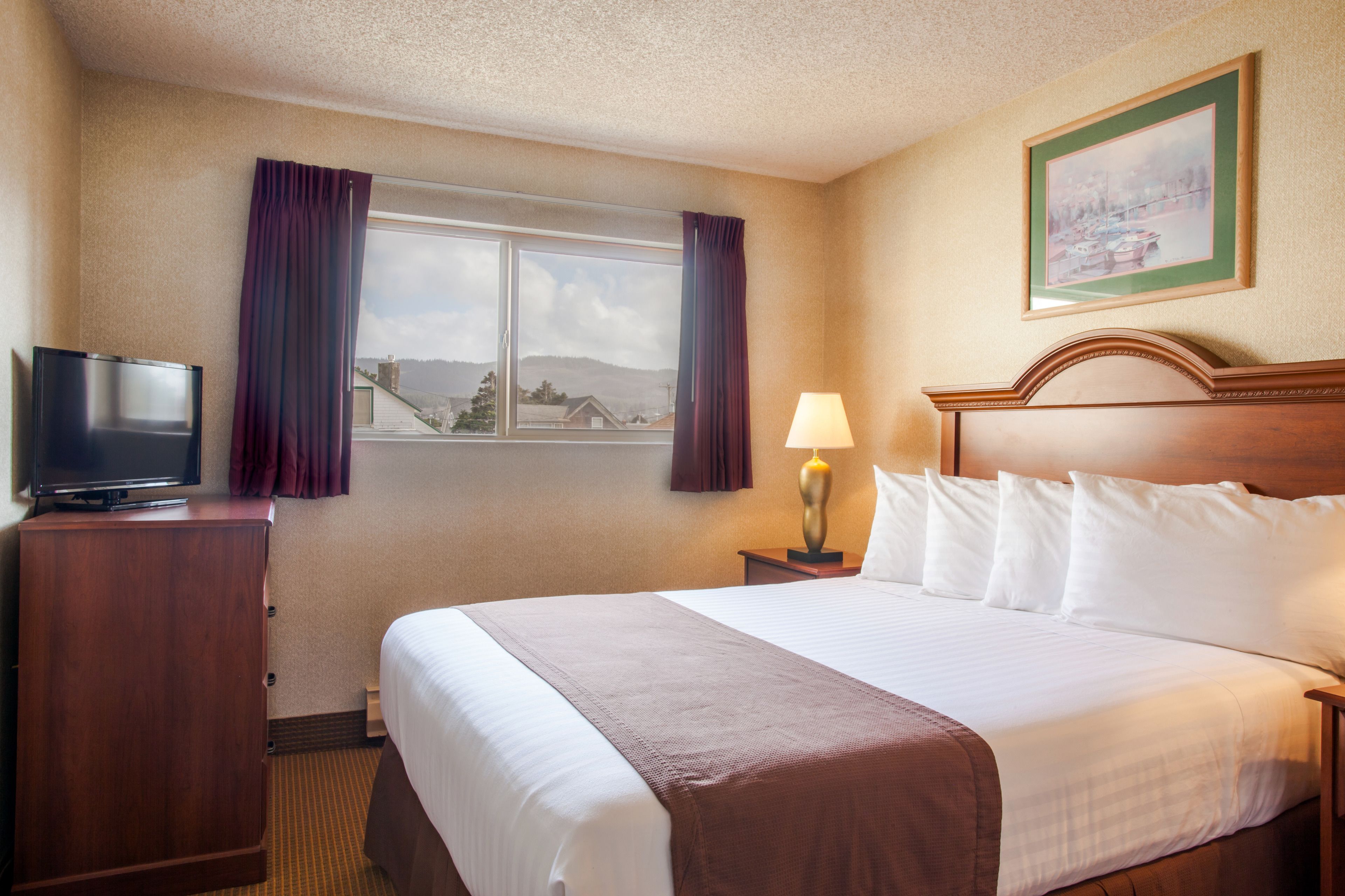 A hotel room with a bed, a TV on a wooden cabinet, a nightstand with a lamp, and a window with curtains.