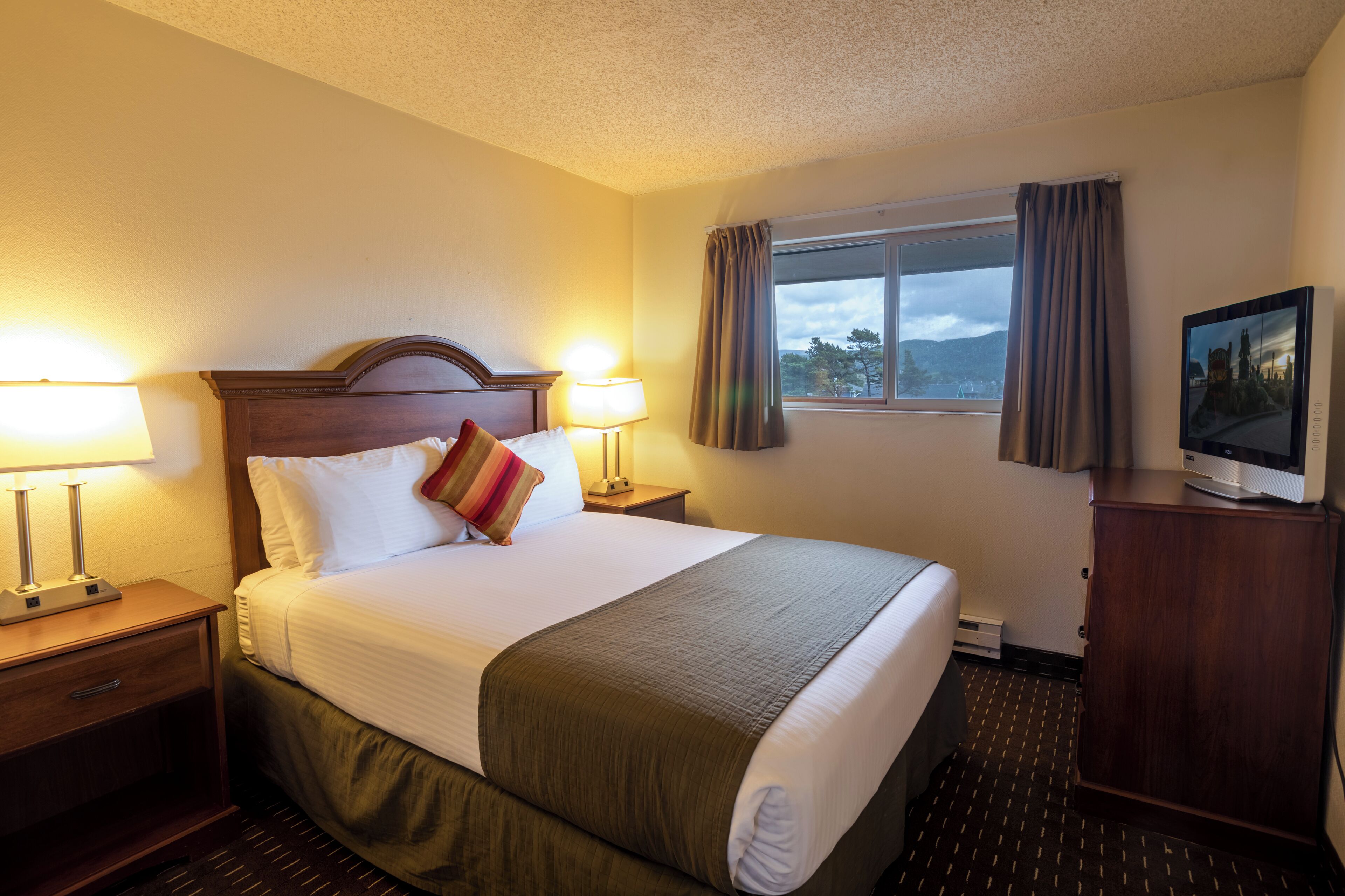 A hotel room with a large bed, two bedside lamps, a television on a wooden cabinet, and a window with curtains.
