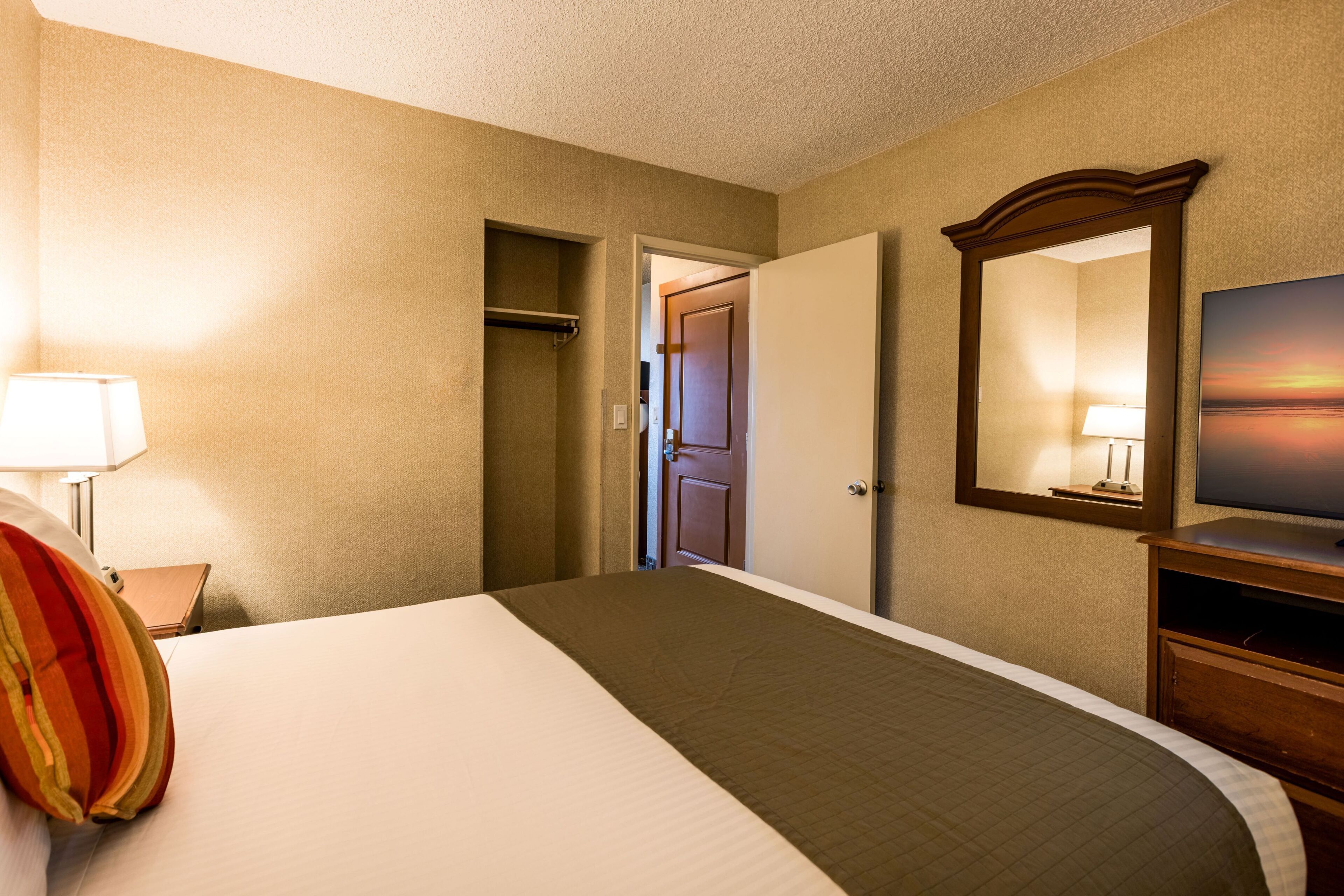 A hotel room with a bed, a dresser, a mirror, a television, and a door leading to another room.