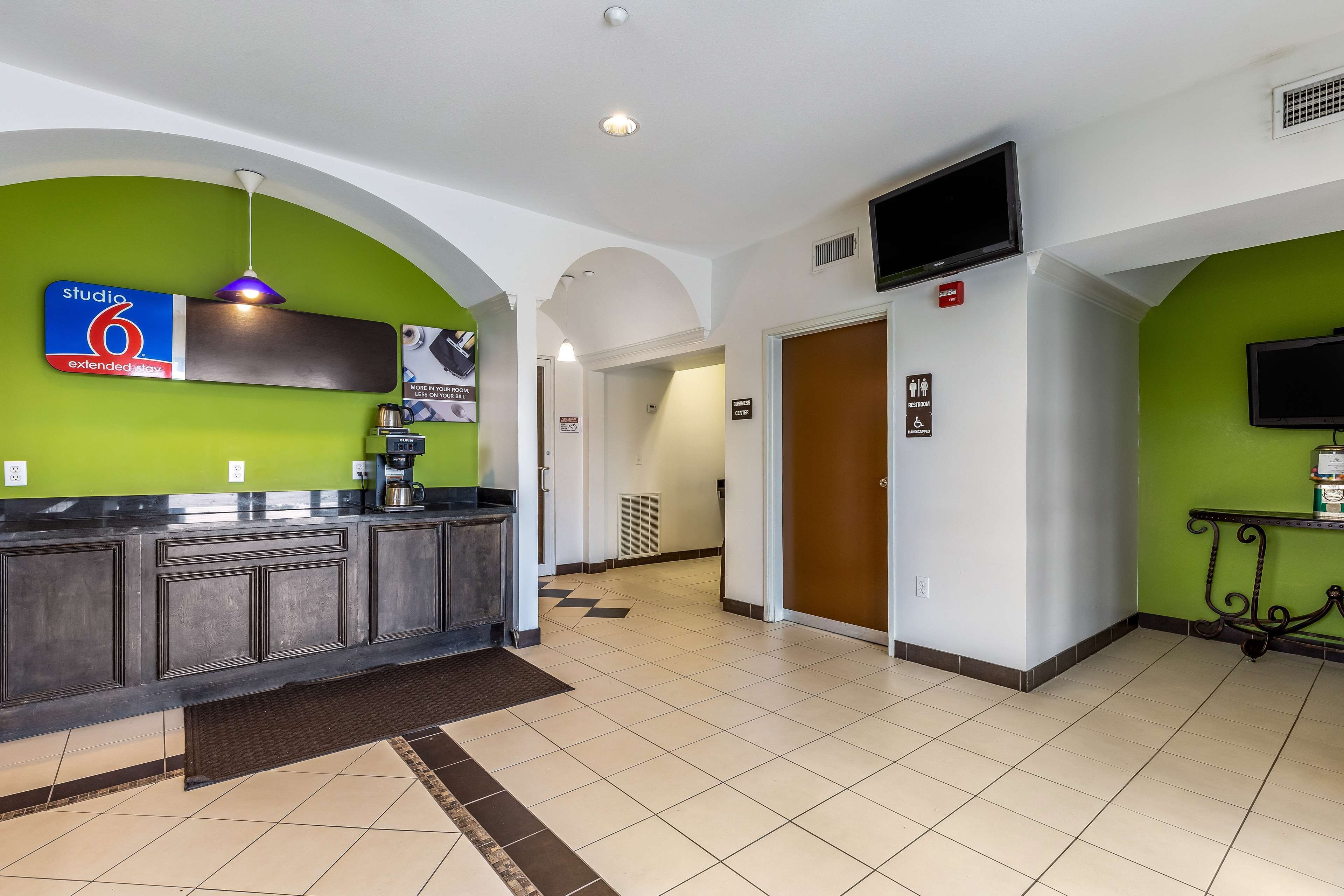 A hotel lobby with a reception desk, a sign for 'Studio 6 Extended Stay', a television mounted on the wall, and a door marked 'Guest Services'.
