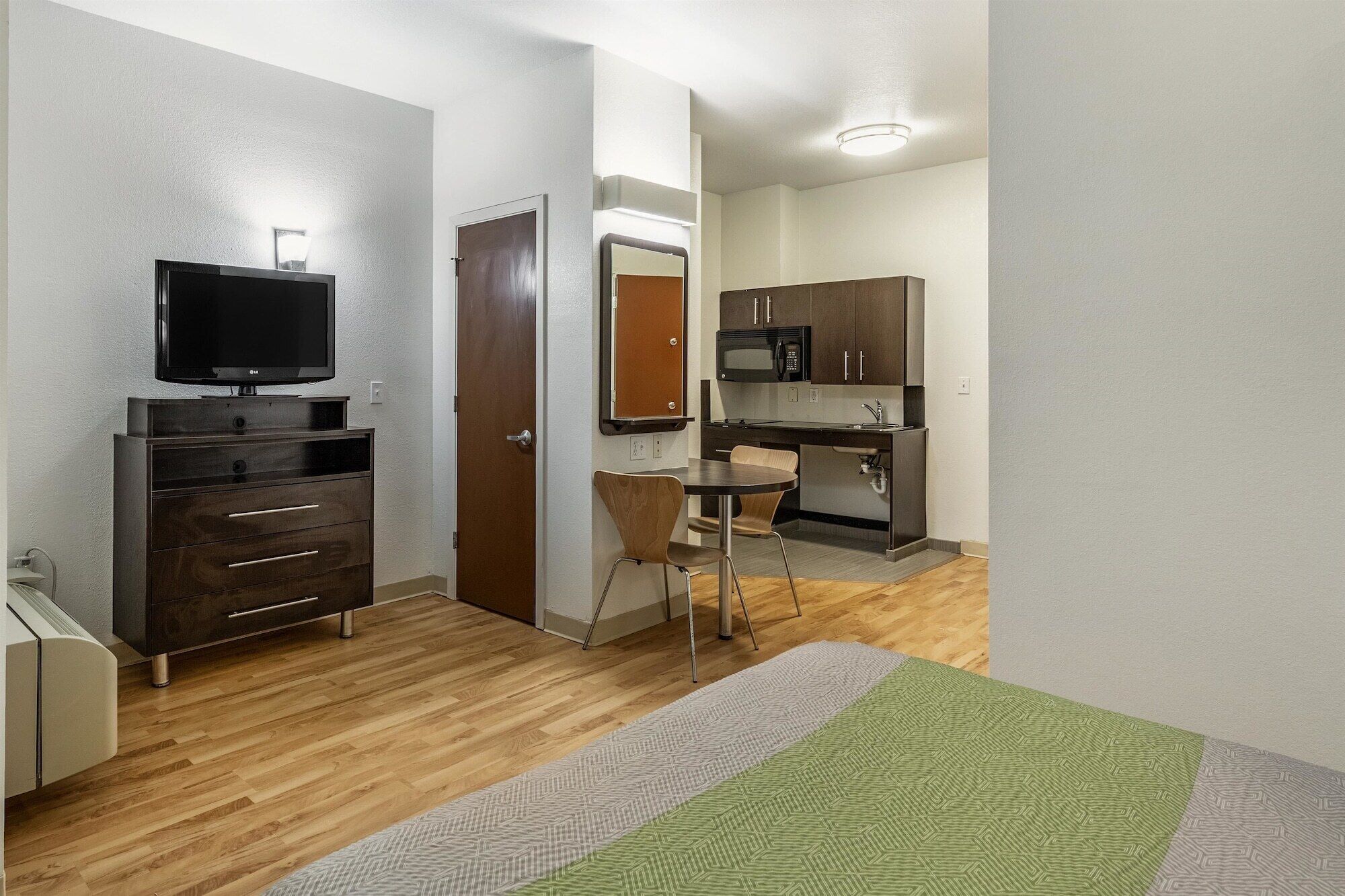 A compact hotel room with a wooden floor, a bed with a green and white patterned bedspread, a small kitchenette with a microwave and dark cabinets, a TV on a stand, and a door leading to a bathroom.