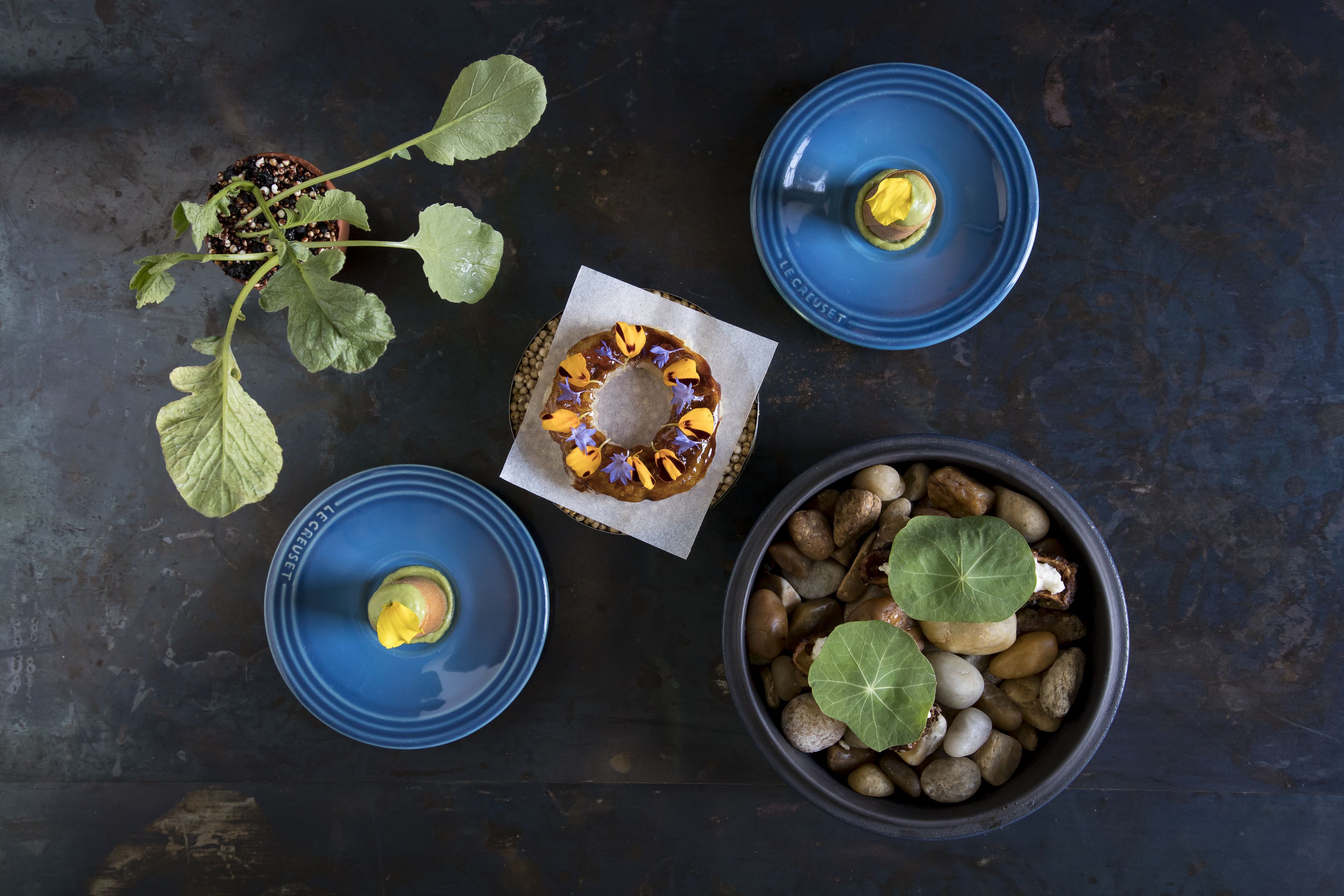 A top-down view of a dark surface with a blue plate containing a yellow object, a blue plate with a yellow object, a bowl with pebbles and a green leaf, and a plant with green leaves and a yellow object.