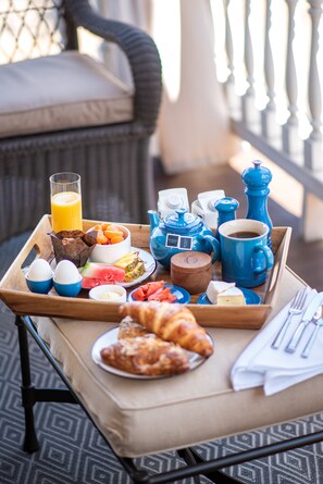 A breakfast tray on a balcony with a wicker chair, a tray with eggs, toast, fruit, coffee, and a croissant.