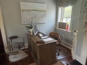 A home office with a desk, computer, and two chairs. There is a window with a view of trees and a painting on the wall.