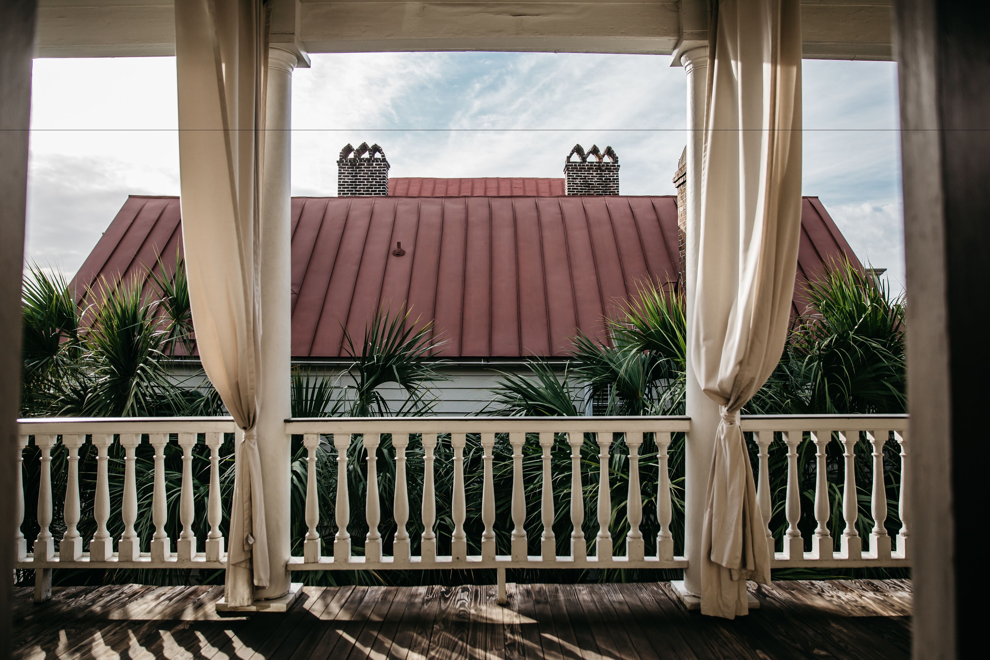 A covered porch with a white balustrade, a red-roofed building in the background, and green foliage.