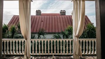 A covered porch with a white balustrade, a red-roofed building in the background, and green foliage.