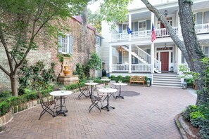 A courtyard with brick flooring, outdoor tables, and chairs, surrounded by greenery and a brick building with a balcony.