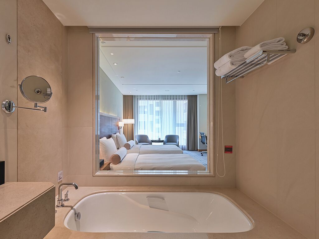 A modern hotel bathroom with a large bathtub, a mirror, and a view of a bedroom with a bed and a desk.