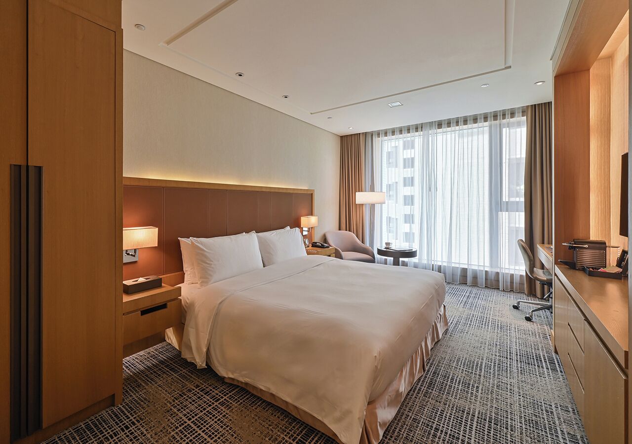 A hotel room with a large bed, a desk, a chair, a lamp, and a view of the city through the window.