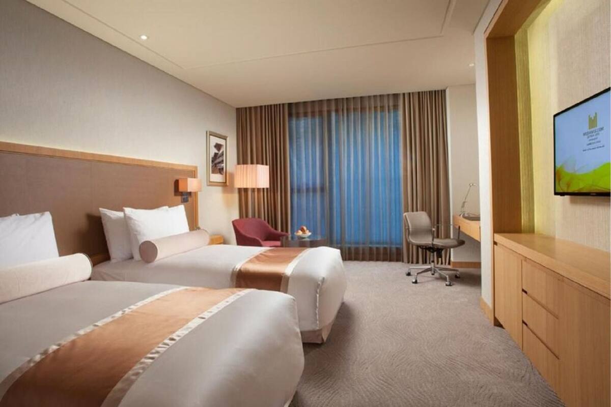 Habitación de hotel con dos camas, un escritorio, una silla, un televisor y un ventanal grande con cortinas.