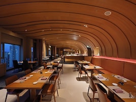 A restaurant with a curved wooden ceiling, long tables with chairs, and a bar area in the background.