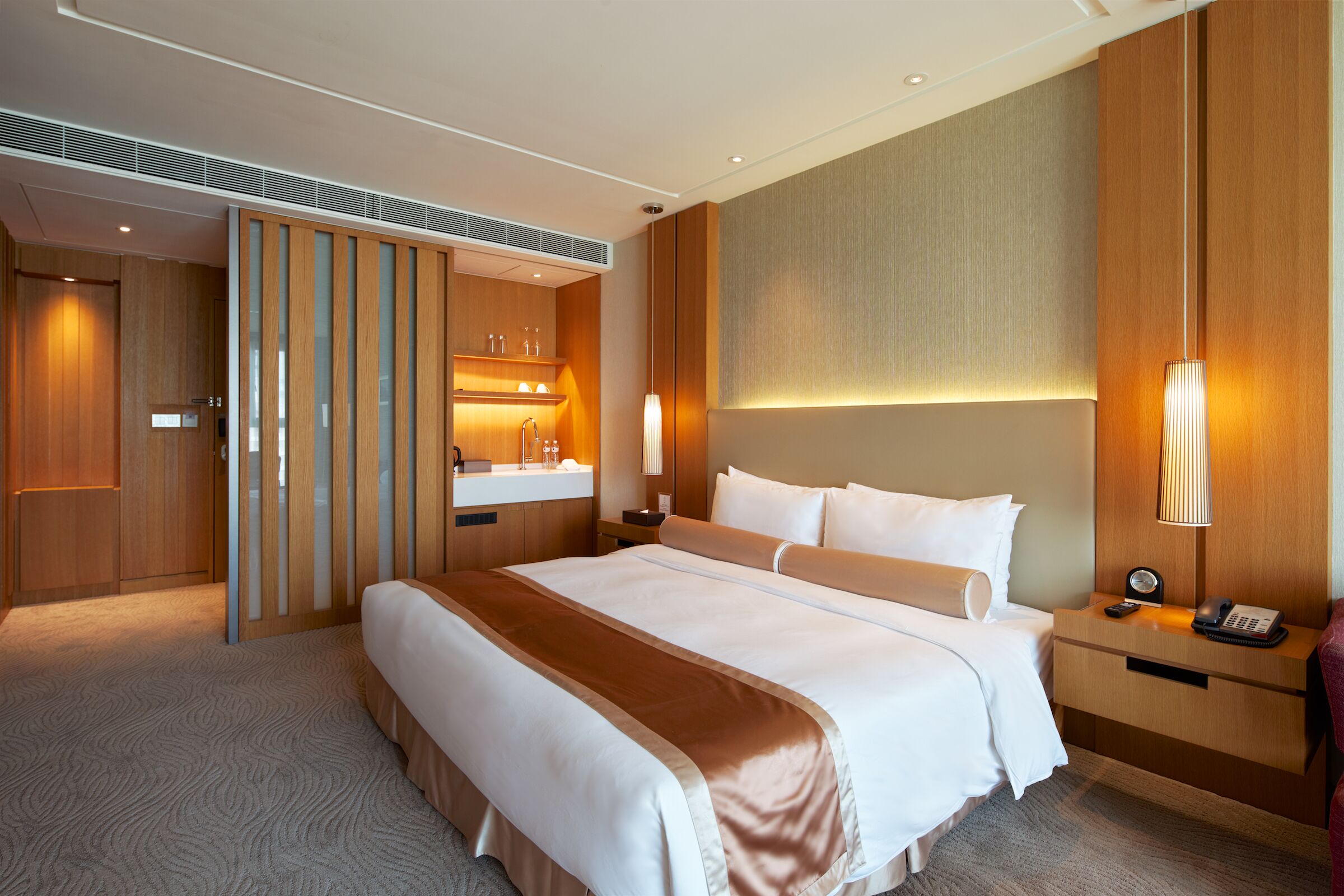 Una habitación de hotel con una cama grande, un armario de madera, una mesita de noche con lámpara y un tocador empotrado con espejo.