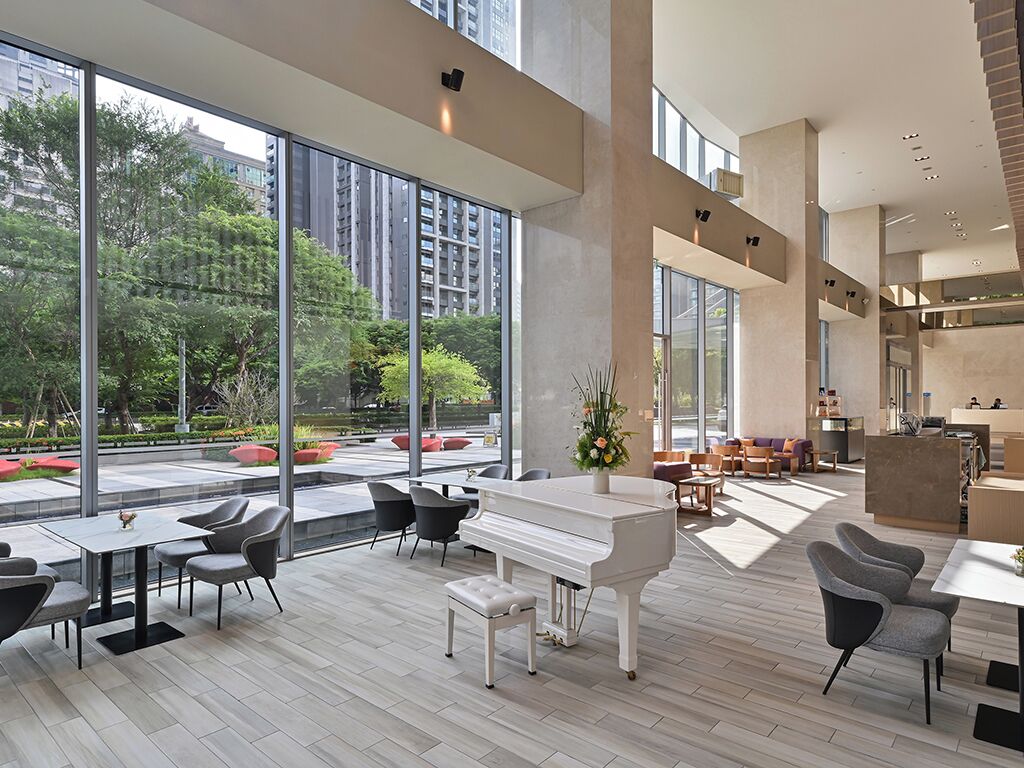 Un espacio interior amplio con un piano, arreglos de asientos y ventanas grandes que ofrecen vistas a zonas verdes y edificios.