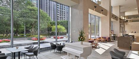 Un espacio interior amplio con un piano, arreglos de asientos y ventanas grandes que ofrecen vistas a zonas verdes y edificios.
