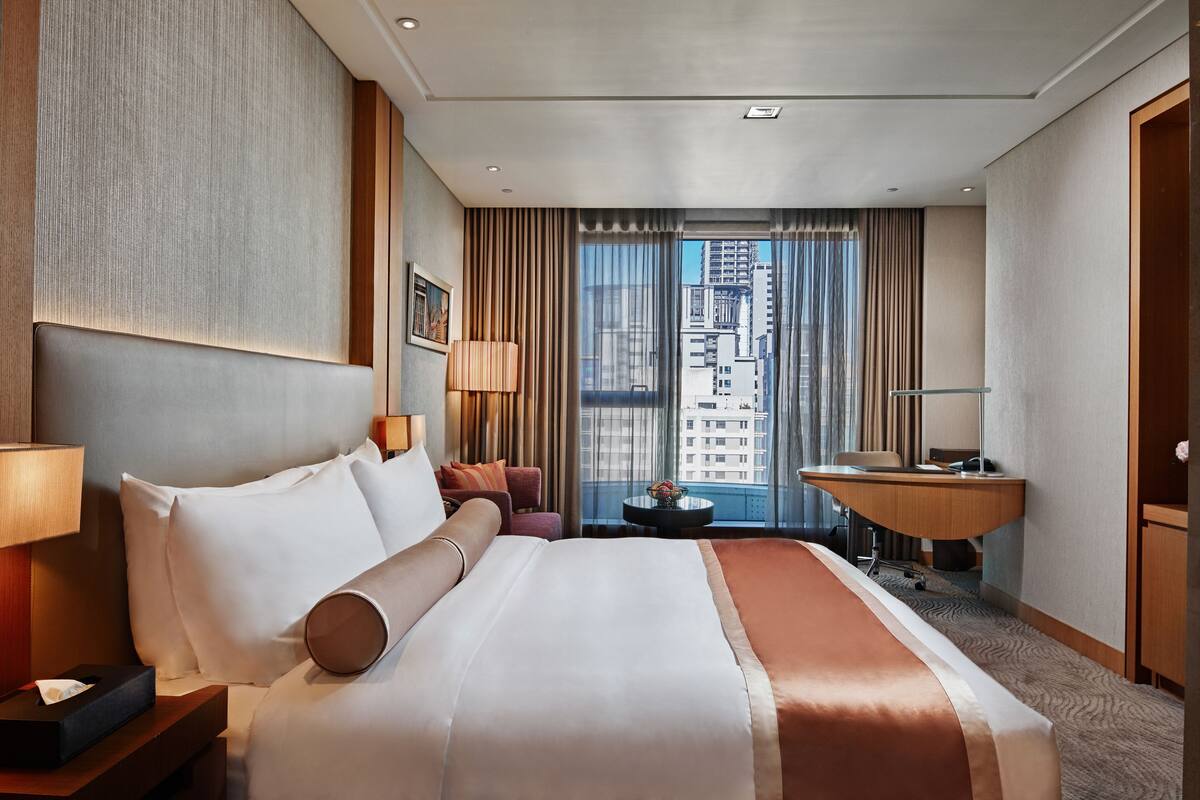 Habitación de hotel con una cama grande, un escritorio, una silla, una lámpara y vistas a la ciudad desde la ventana.