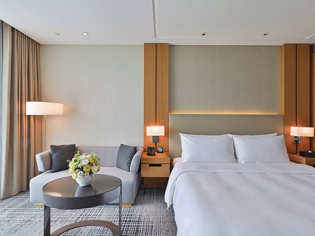Une chambre d’hôtel avec un lit, un canapé, une table ronde ornée d’un vase de fleurs, deux lampes de chevet et une grande fenêtre avec des rideaux.