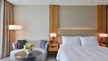 A hotel room with a bed, a sofa, a round table with a vase of flowers, two bedside lamps, and a large window with curtains.