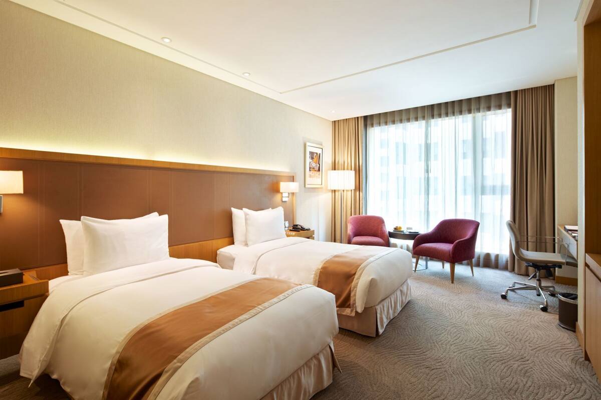 Habitación de hotel con dos camas, un escritorio, una silla y un ventanal con cortinas.