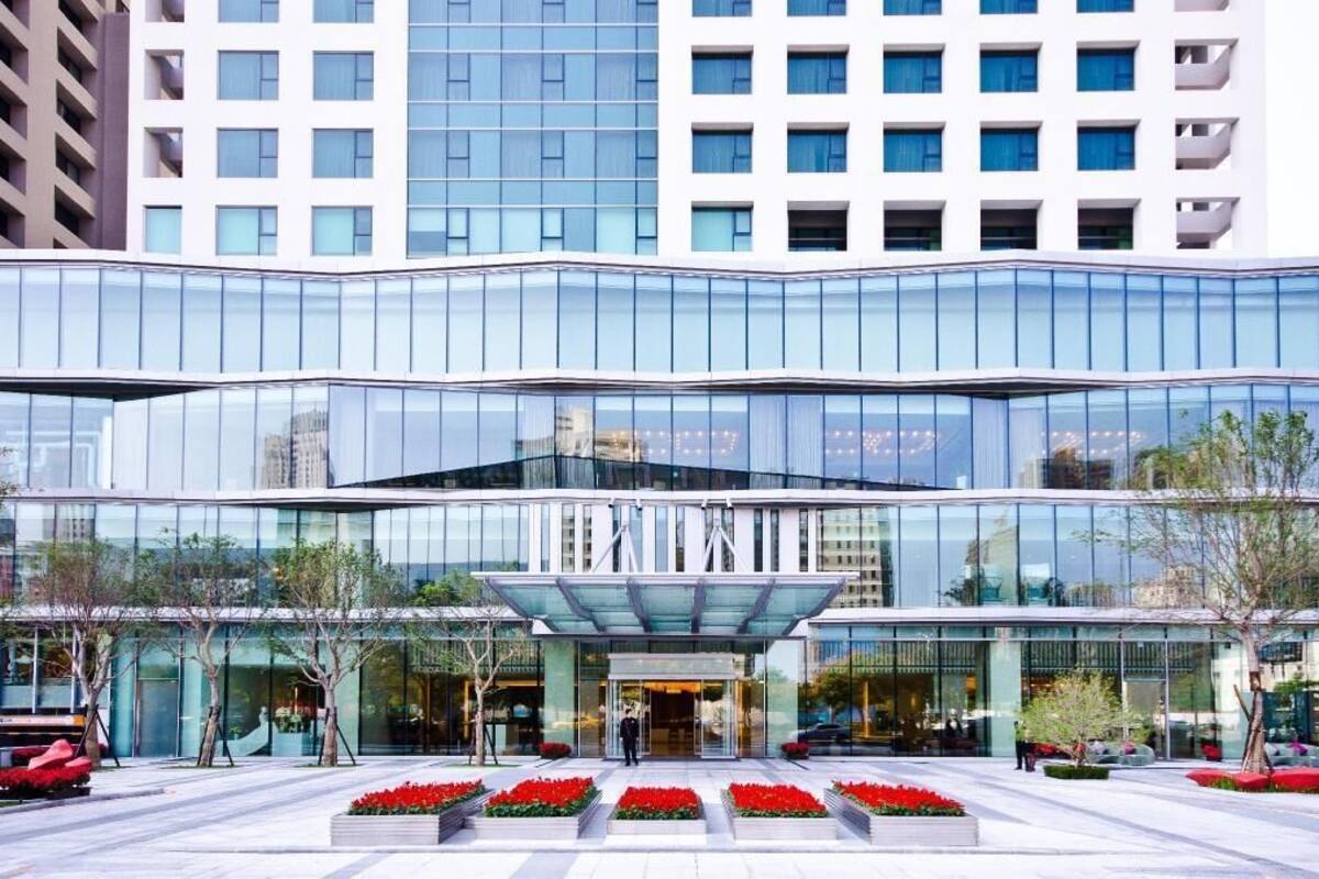 Un hotel moderno con fachada de vidrio, entrada central y área ajardinada con flores rojas.