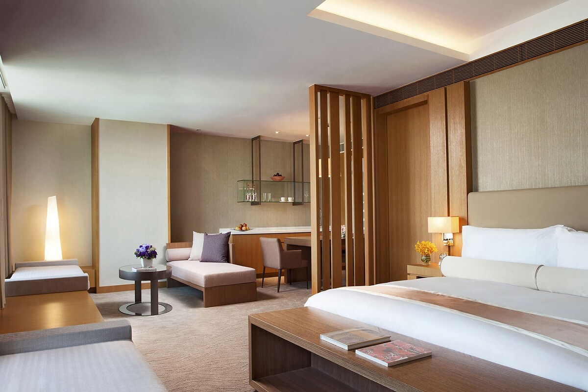 Una habitación de hotel moderna con una cama grande, un escritorio, una silla, una mesita y un espacio de baño con espejo y estanterías.