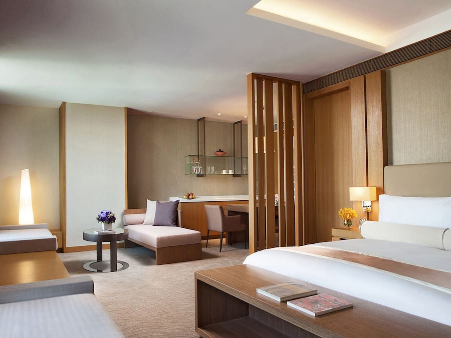 A modern hotel room with a large bed, a desk, a chair, a small table, and a bathroom area with a mirror and shelves.