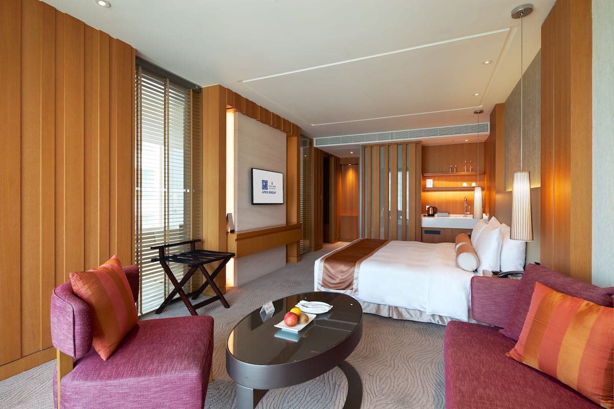 Habitación de hotel con cama, dos sillones, una mesita con una bandeja de frutas, un escritorio con silla y baño visible a través de una puerta corredera.