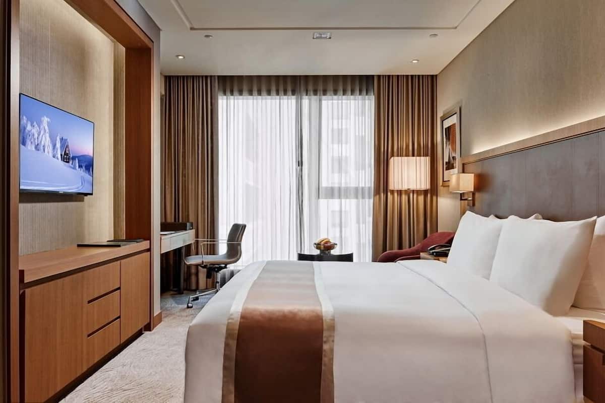 Habitación de hotel con una cama grande, un escritorio con silla, un televisor en la pared y una ventana con cortinas.