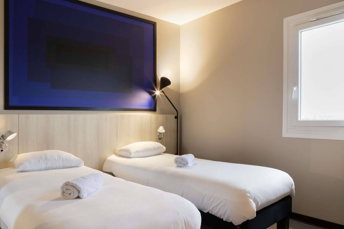 Habitación de hotel con dos camas, un cuadro abstracto grande, una lámpara de pie y una ventana.