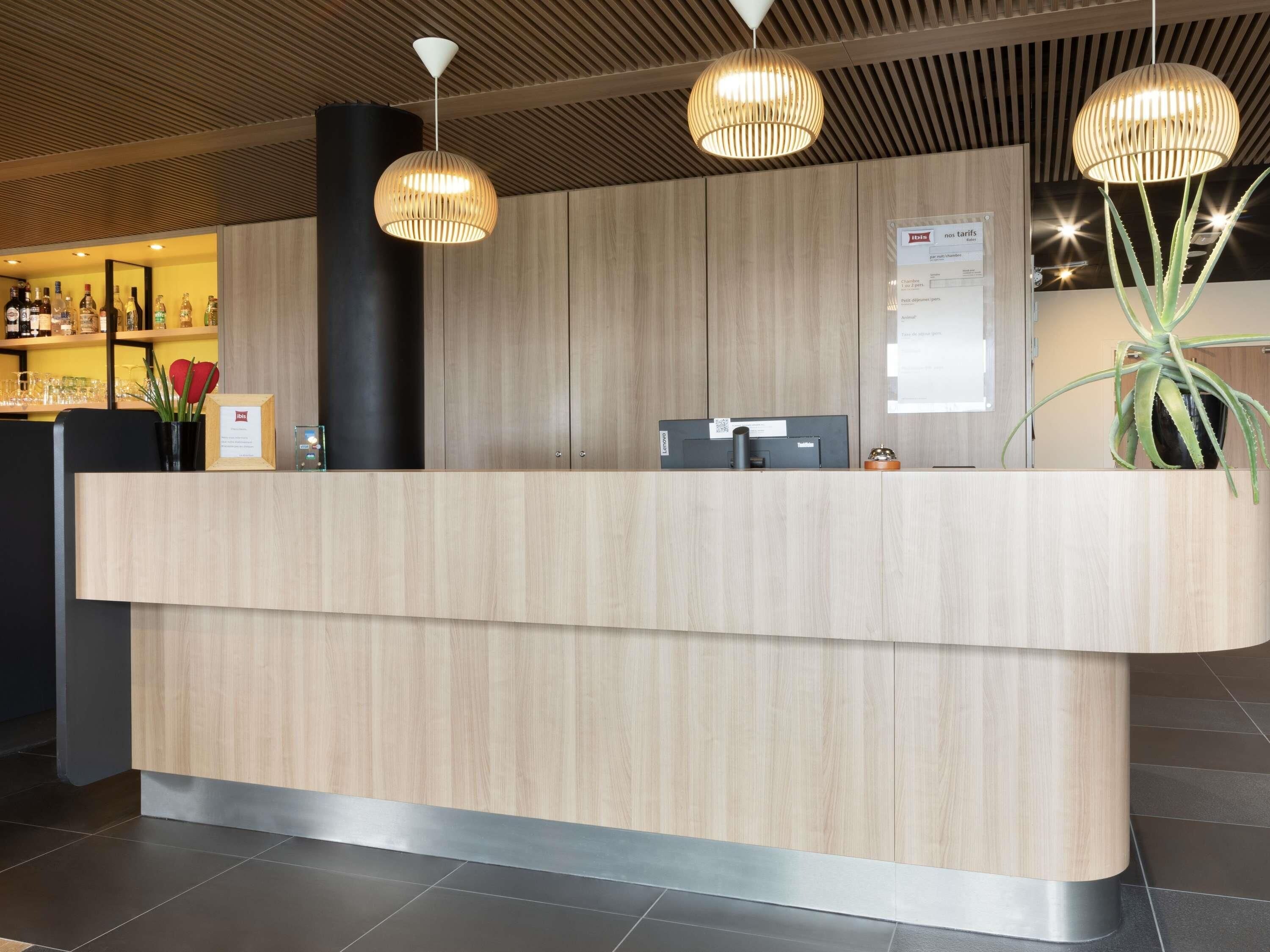 A reception desk with a wooden finish, a black pillar, and a digital screen. There are two pendant lights above the desk, and a potted plant to the right.