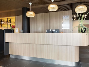 A reception desk with a wooden finish, a black pillar, and a digital screen. There are two pendant lights above the desk, and a potted plant to the right.
