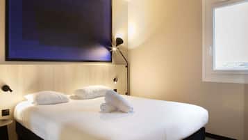 A neatly made bed with white linens and two pillows, a wall-mounted TV, a bedside lamp, and a window with a view.