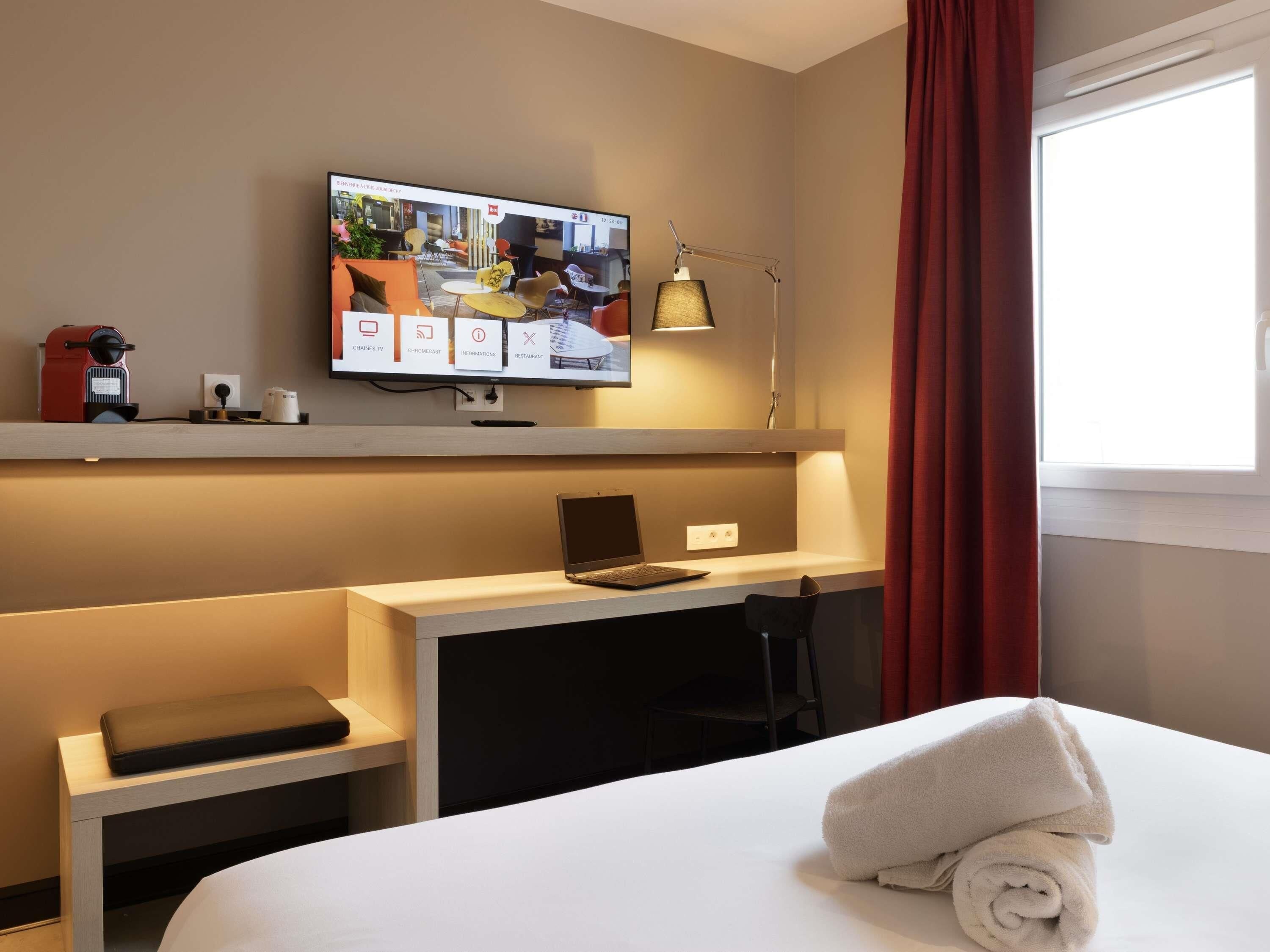 Une chambre d’hôtel avec un lit, un bureau, un ordinateur portable, un téléviseur fixé au mur et une fenêtre avec des rideaux.