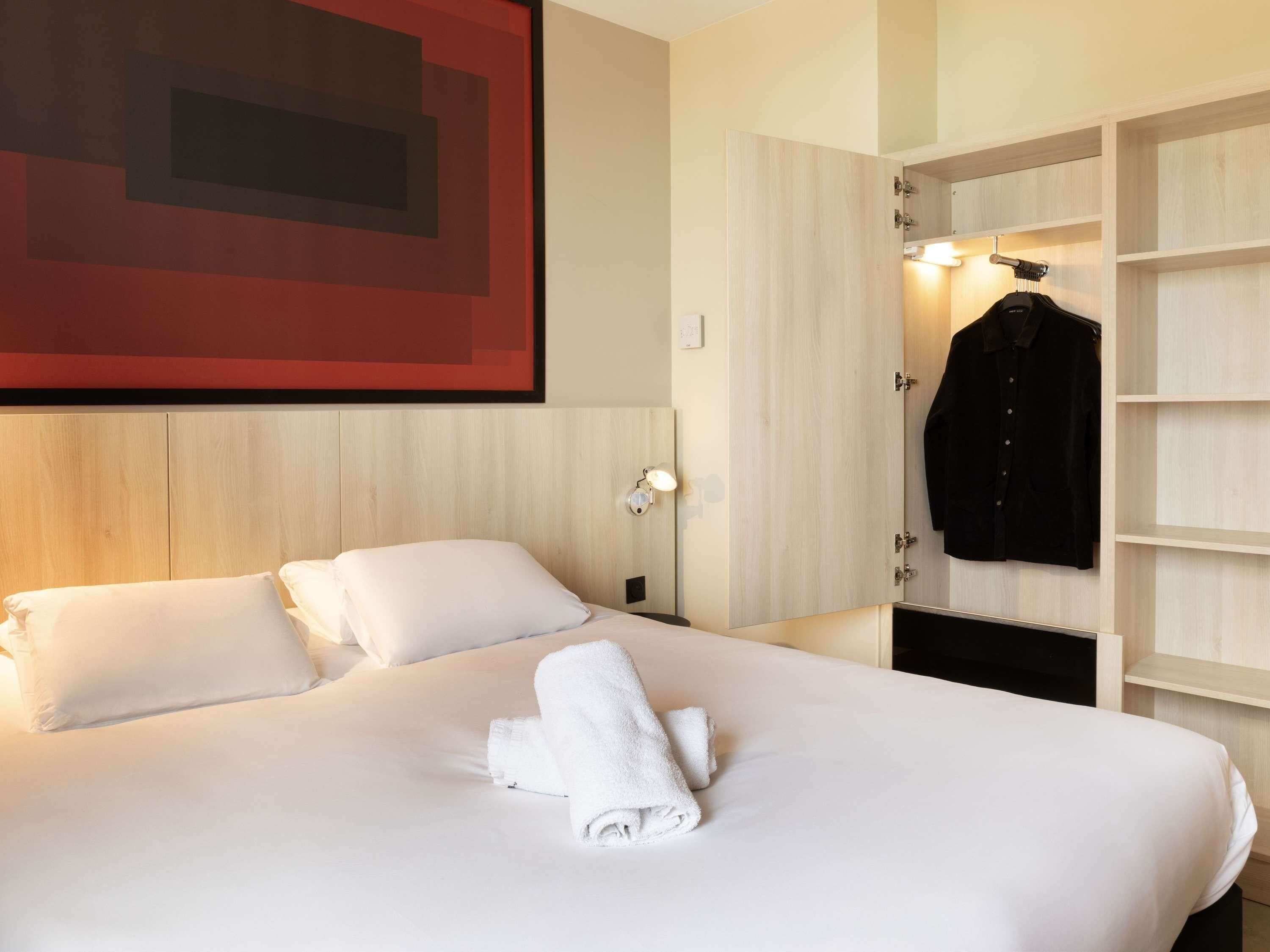 Habitación de hotel con cama, ropa de cama blanca, toalla doblada con cuidado, abrigo colgado y una lámpara de pared.