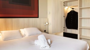 A hotel room with a bed, white linens, a neatly folded towel, a hanging coat, and a wall-mounted light. - ibis Douai Dechy (Dechy)