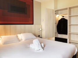 Habitación de hotel con cama, ropa de cama blanca, toalla doblada con cuidado, abrigo colgado y una lámpara de pared.