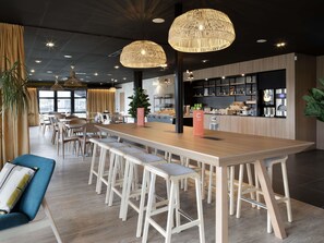 Un comedor moderno con una larga mesa de madera, taburetes de bar, iluminación colgante y una cocina al fondo.