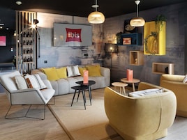 Un intérieur moderne comprenant un canapé, des fauteuils, une table basse, une bibliothèque et un téléviseur fixé au mur, sur lequel est affiché le logo d’Ibis.