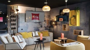 A modern interior with a sofa, armchairs, a coffee table, a bookshelf, and a mounted television displaying the ibis logo. - ibis Douai Dechy (Dechy)