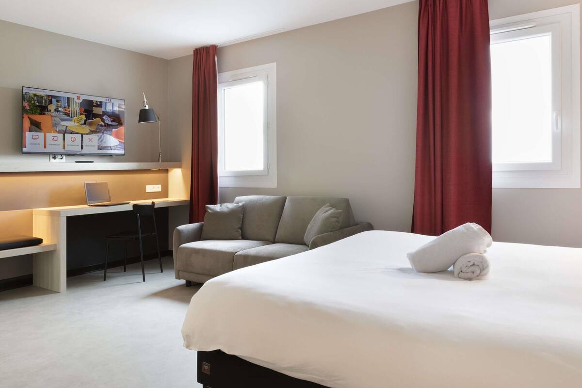 Una habitación de hotel con cama, escritorio, silla, televisor y cortinas rojas.