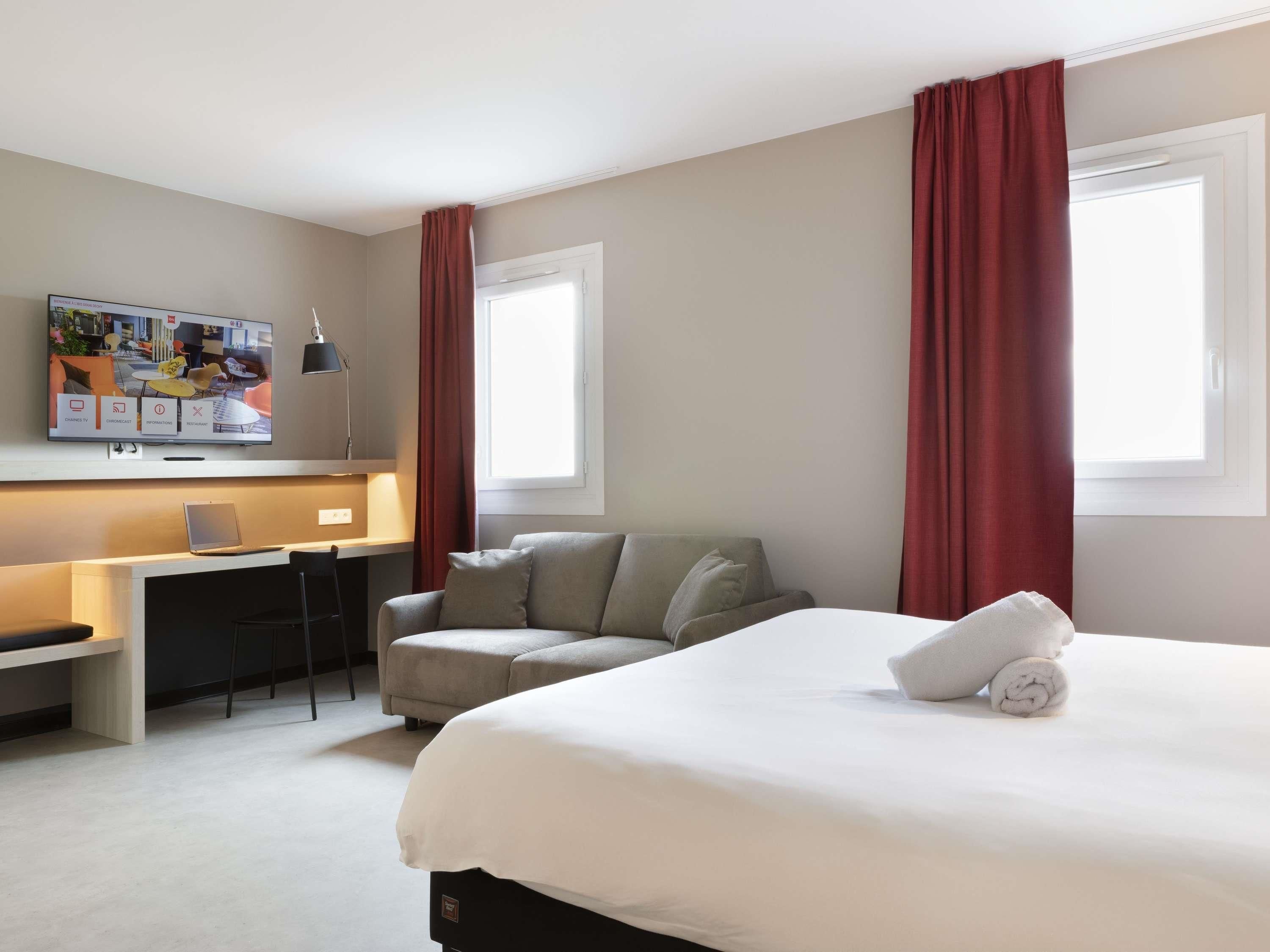 Habitació d'hotel amb un llit, una taula de treball, una cadira, un televisor i cortines vermelles.