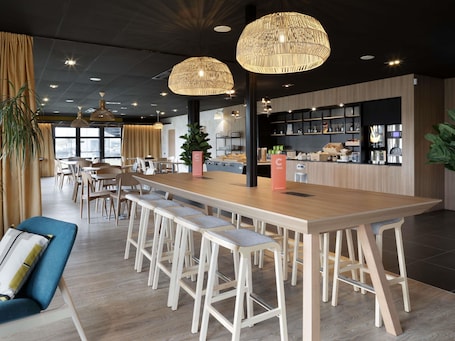 A modern dining area with a long wooden table, bar stools, pendant lighting, and a kitchen in the background.. ibis Douai Dechy
