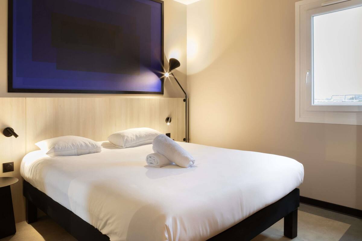 Una cama bien tendida con sábanas blancas y dos almohadas, un televisor montado en la pared, una lámpara de noche y una ventana con vistas.