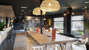 A modern cafe with a long wooden table, bar stools, and hanging wicker lamps. - ibis Douai Dechy (Dechy)