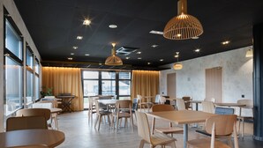 A modern dining area with wooden tables and chairs, pendant lighting, and large windows allowing natural light. - ibis Douai Dechy (Dechy)