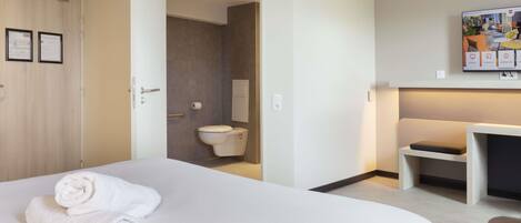 Habitación de hotel con una cama grande, una mesita de noche, una silla, un escritorio, un inodoro y un televisor montado en la pared.