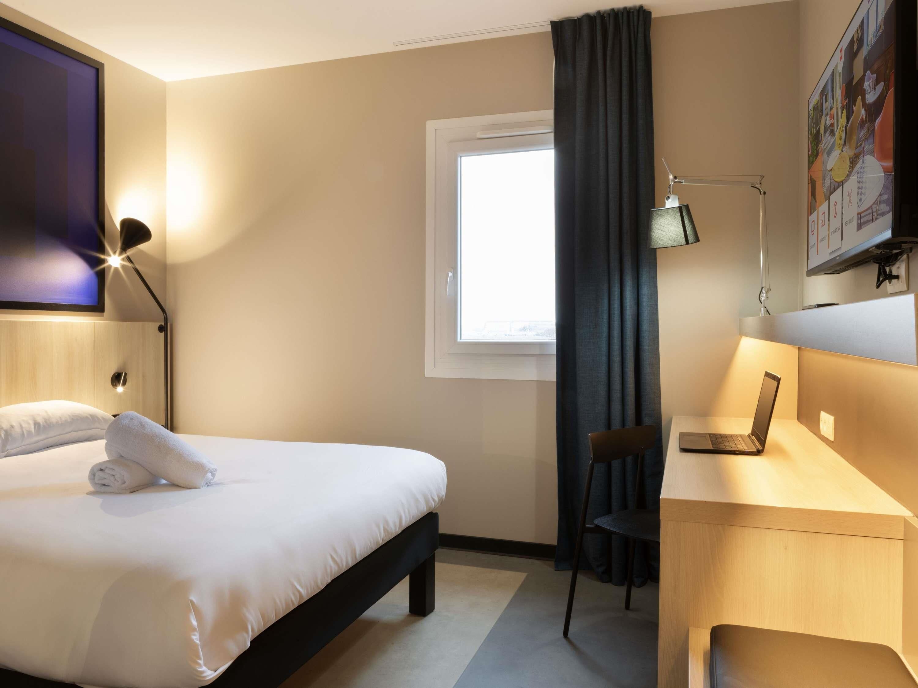 Habitació d'hotel amb un llit, una taula de treball amb un ordinador portàtil, una cadira, una finestra amb cortines i una televisió penjada a la paret.