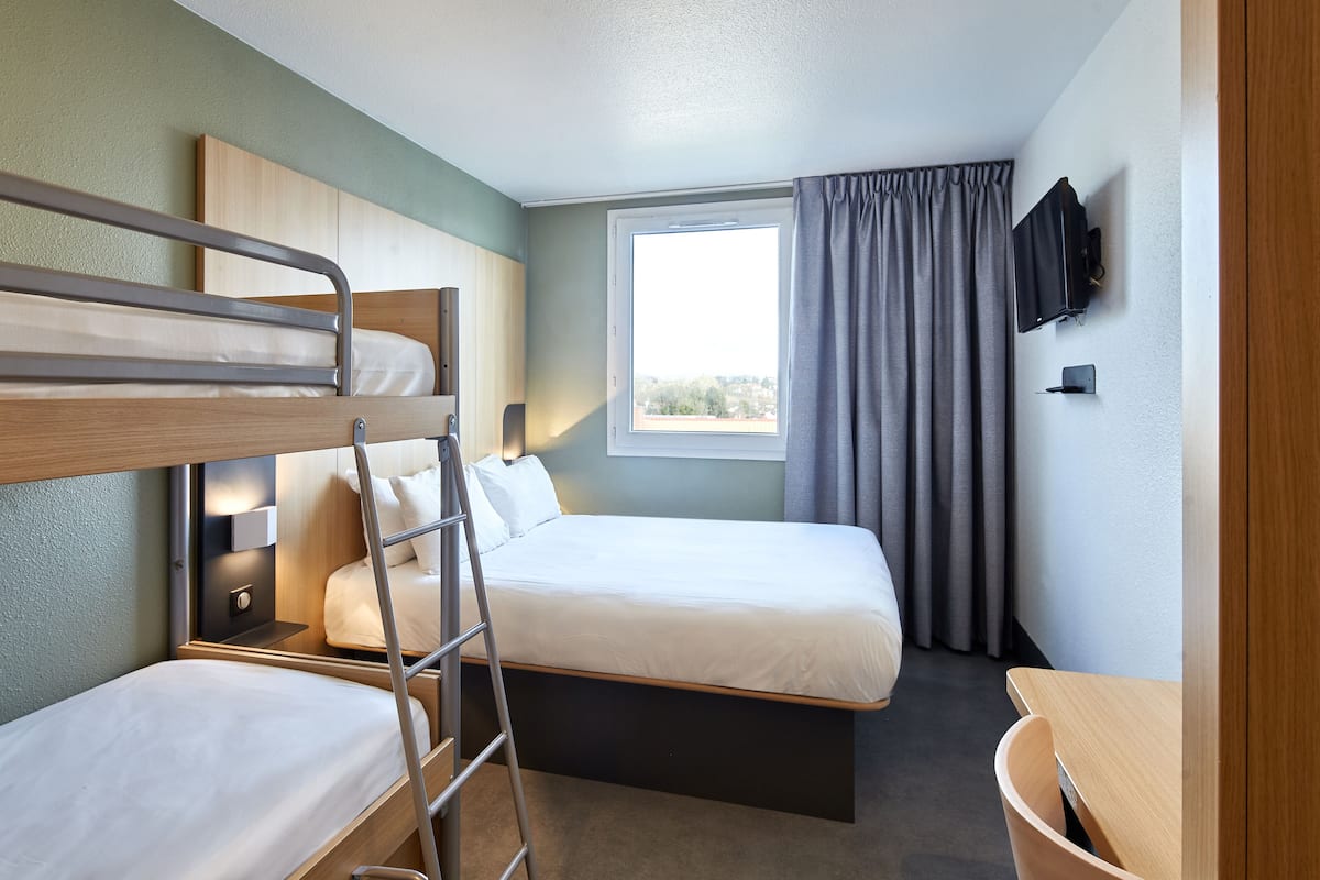 Une chambre d’hôtel avec un lit superposé, un lit simple, un bureau, une chaise, une télévision et une fenêtre avec des rideaux.